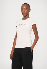 Tricou fitted de culoare roz deschis, cu guler rotund și mâneci scurte, având un logo din text reliefat, asortat cu pantaloni negri care prezintă accente aurii.