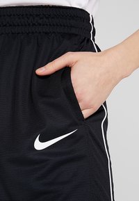 Pantalones cortos deportivos negros con una tela texturizada, con franjas laterales blancas y el logo de Nike en forma de lápiz, mostrando una mano en el bolsillo lateral.
