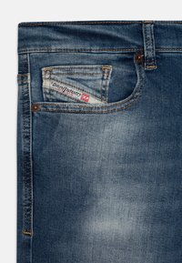 Jeans de denim em um tom de azul médio com desbotamento leve, apresentando um bolso frontal com um rótulo vermelho e branco, e detalhes de costura visíveis.