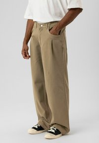 Beige wijde broek met plooien aan de voorkant, zijzakken en een knoopsluiting. Gecombineerd met zwart-witte sneakers. Glad stoftextuur.