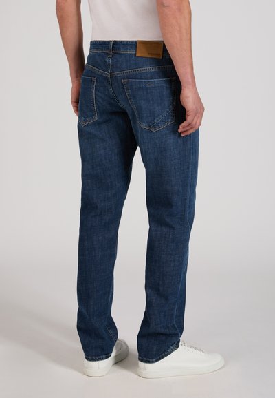 Man wearing dark blue jeans and white sneakers, standing with back facing the camera against a plain background.