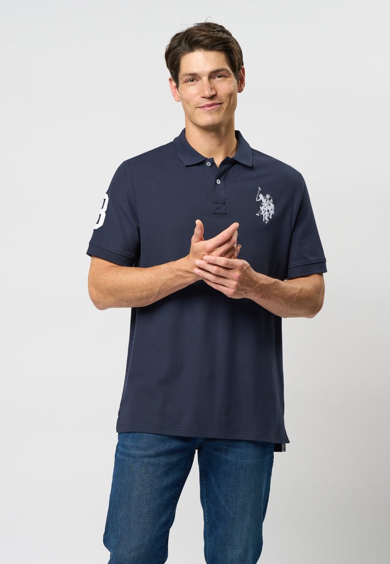 Námořní polo tričko so golierom, trojicoch gombíkmi a krátkymi rukávmi. Biele logo a veľký detail čísla na ľavom rukáve.