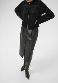 Person in einem schwarzen, gefransten Reißverschluss-Pullover, schwarzer, durchbrochener Leder-Midi-Rock mit vorderem Schlitz und schwarzen spitzen Stiefeln, steht mit den Händen in den Hüften.