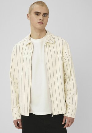 Veste de couleur crème avec des rayures verticales noires, fermeture éclair, col pointu et manches longues, portée sur une chemise blanche.