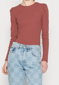 Rostfarbener Langarmoberteil mit ausladendem Schulterdetail, kombiniert mit hellblauen karierten High-Waist-Jeans, die einen Reißverschluss und einen Knopfverschluss haben.