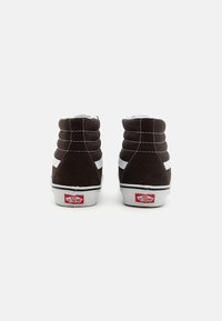 Tmavohnedé vysoké tenisky so bielymi akcentmi, mäkký semišový materiál, viditeľné švy a logo Vans na päte. Okrúhly dizajn špičky.