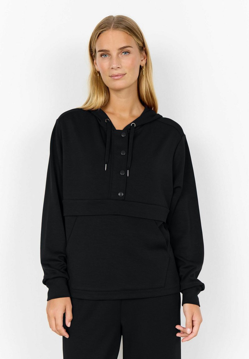 Jeune femme avec de longs cheveux blonds portant un sweat à capuche noir avec des détails de boutons et un pantalon noir assorti, se tenant devant un fond blanc.