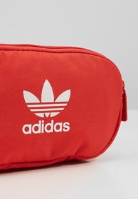 Sacchetto di tessuto rosso con una superficie liscia, caratterizzato da un logo Adidas bianco e un design con tre strisce. Chiusura con zip nella parte superiore.