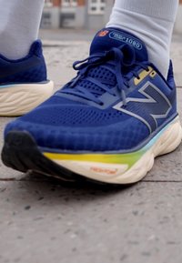 Chaussure de course bleue avec une texture en nid d'abeille, dotée d'un logo gris sur le côté, d'une semelle blanche avec des accents jaunes et verts, et d'une semelle extérieure sombre.