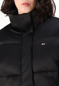 Czarna kurtka puchowa z wysokim kołnierzem, pikowanym wzorem i subtelnym logo na piersi. Materiał wydaje się gładki i błyszczący.