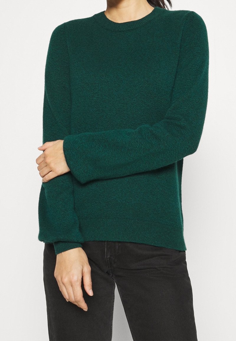 Personne portant un pull en maille à manches longues vert foncé et un pantalon noir, les bras légèrement croisés à la taille.