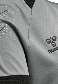 Harmaa urheilu-paita, jossa on tekstuuroitu pinta, graafinen kuviot ja musta verkkomallinen kaulus. Logo on painettu tummanharmaalla.