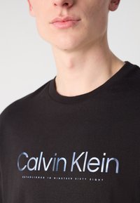 Sort t-shirt lavet af bomuld, med et trykt "Calvin Klein" logo i metallisk gradient og tekst nedenfor i hvid, rund halsudskæring.