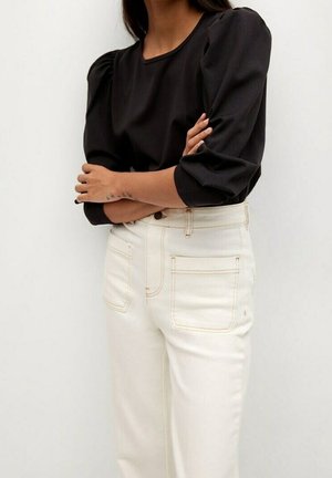 Vrouw draagt een zwarte blouse met pofmouwen en een hoog uitgesneden, off-white broek met frontzakken, armen gekruist.