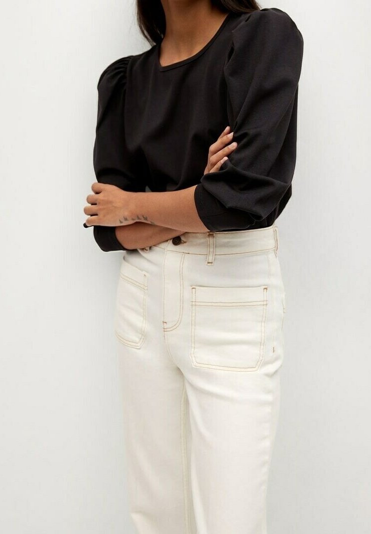 Mujer con blusa negra de mangas abullonadas y pantalones de talle alto color blanco roto con bolsillos parche delanteros, brazos cruzados.