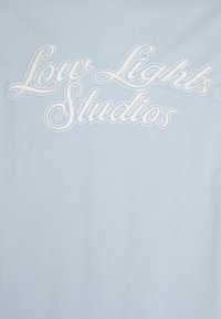Světle modré bavlněné tričko s odvážnou bílou křivkovou výšivkou se nápisem "Low Lights Studios" na přední straně. Hladký povrch.