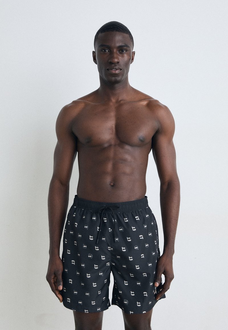 Sorte badeshorts med et gentaget grafisk mønster, elastisk talje og snøre detaljer, båret af en shirtløs mandlig model.