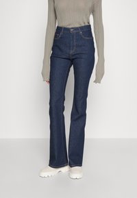 Dunkelblaue Flared-Jeans aus Denim mit kontrastierenden Nähten. Kombiniert mit einem hellgrauen, gerippten Langarmoberteil und beigen Plateauschuhen.