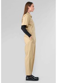 Combinaison utilitaire beige à manches courtes, avec deux poches latérales et une coupe décontractée. Portée par-dessus un haut à manches longues noir en tissu texturé.