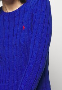 Blå kabelstickad tröja med rund halsringning och ribbade muddar, med en liten röd polo-logga på bröstet.