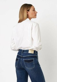 Woman with straight blonde hair wearing a white button-up shirt and dark blue jeans with studded back pockets, facing sideways.