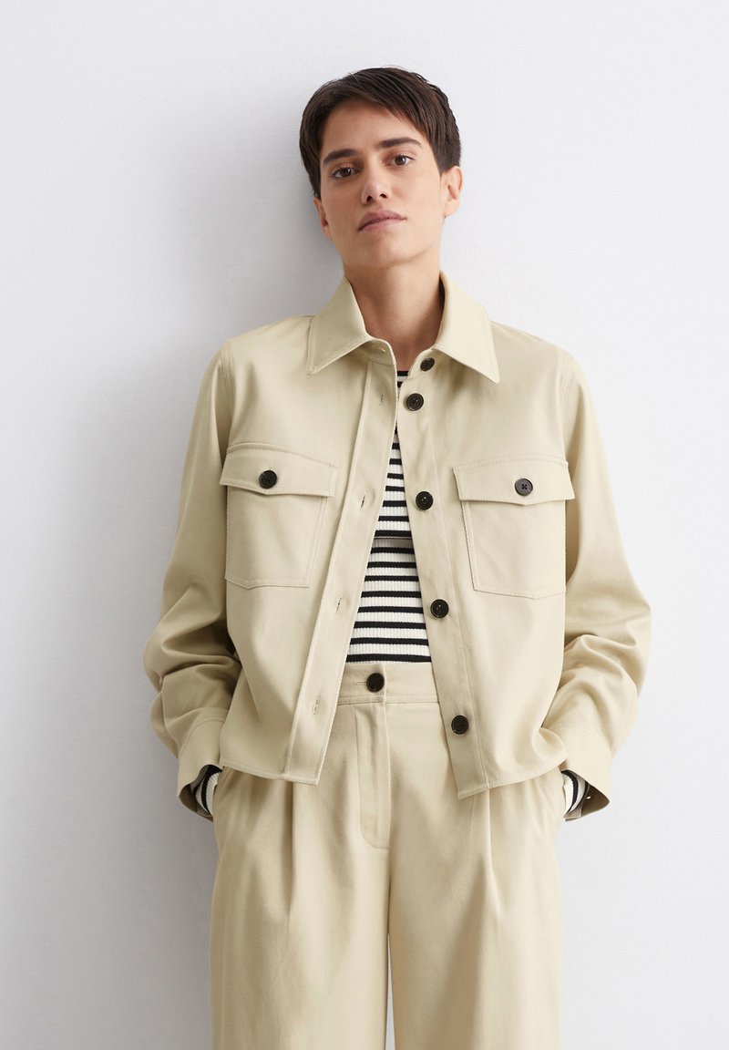 Beige Jacke mit zwei geknöpften Brusttaschen, einem spitzen Kragen, kombiniert mit einem gestreiften Oberteil und weitgeschnittenen beigen Hosen. Glatte Stofftextur.