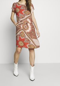 Czerwona sukienka w drobny wzór paisley z krótkim rękawem, luźnym krojem i bocznymi kieszeniami. Wzór zawiera elementy kwiatowe i geometryczne. Noszona z białymi botkami na kostkę.