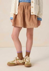 Enfant portant une jupe en velours côtelé marron, des chaussettes marron avec des cœurs roses et un bord violet, des baskets beiges, un t-shirt bleu et un cardigan en tricot crème.