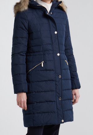 Femme portant un long manteau d’hiver matelassé bleu marine avec capuche bordée de fourrure, poches zippées et boutons-pression argentés sur un pull blanc.