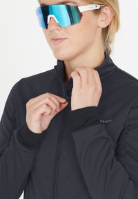 Svart zip-up jacka med hög krage, med en texturerad tyg. Modellen bär stora reflekterande tealfärgade solglasögon.