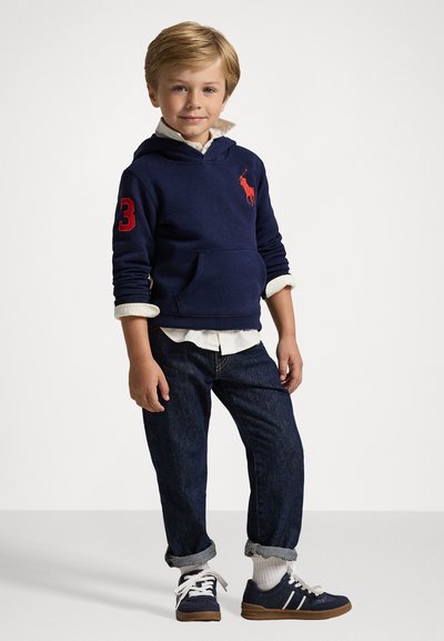 Sudadera marina con logotipo rojo y el número "3" en la manga, camiseta blanca con cuello debajo, jeans oscuros ajustados, zapatillas marinas con acentos blancos.