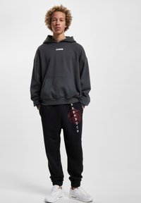 Grauer Hoodie mit Fronttasche und weißem Logo; schwarze Jogginghose mit ro Grafiken und weißem Text; kombiniert mit weißen Sneakern.