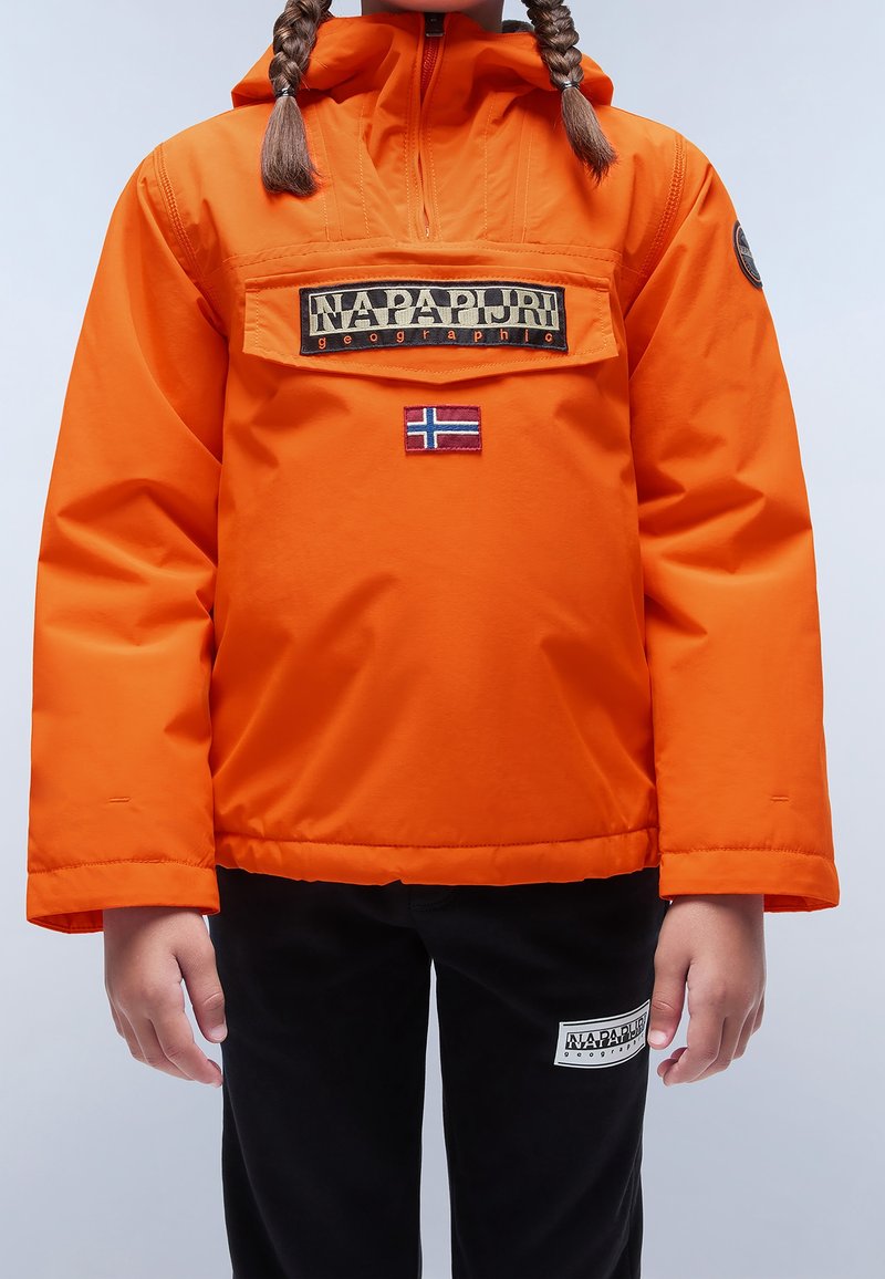 Enfant portant une veste orange vif Napapijri avec une poche avant et un écusson du drapeau norvégien, associée à un pantalon noir avec un écusson logo.