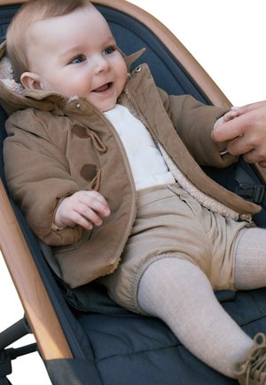 Bambino sorridente con giacca e pantaloni beige sdraiato in un passeggino, che tiene delicatamente la mano di un adulto, vestito in modo caldo per il tempo fresco.