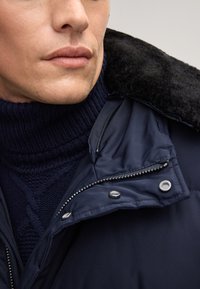 Giacca imbottita blu scuro con un morbido colletto in shearling black, dotata di una chiusura lampo frontale e di un dolcevita in maglia blu navy sotto.
