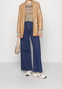 Brązowa skórzana dwurzędowa marynarka na beżowym i brązowym swetrze w wzory, połączona z wysokim stanem szerokimi niebieskimi dżinsami i białymi sneakrami.