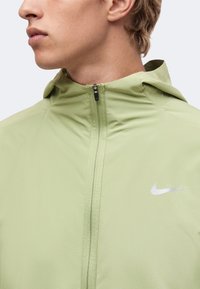 Jeune personne portant une veste à capuche zippée Nike vert clair, montrant le profil latéral du visage et du haut du corps.