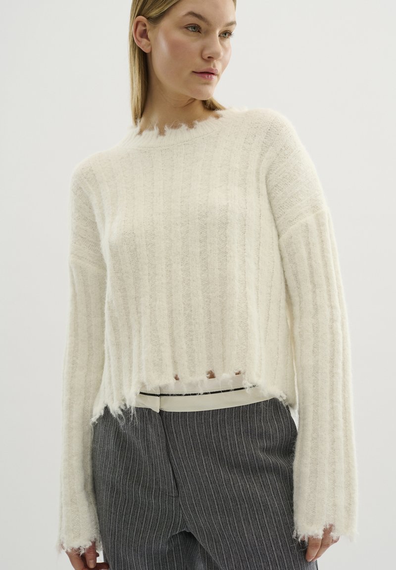 Leichter, off-white gestrickter Pullover mit vertikalen Rippenmustern, fransigem Saum und langen Ärmeln. Kombiniert mit grauen Nadelstreifen-Hosen und einem weißen Gürtel.