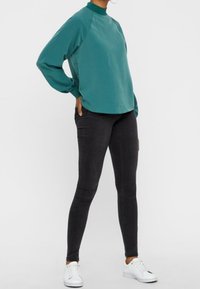 Camicia oversize color teal con collo alto e maniche raglan abbinata a jeans skinny neri e sneakers bianche. Tessuto liscio con dettagli minimi.