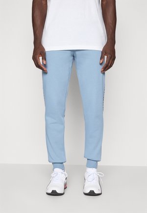 Lyseblå sweatpants med tætsiddende pasform, ribbede manchetter og sidelister, parret med hvide sportssko. Glat overflade, afslappet design.