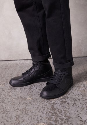 Sorte høje sneakers med snørebånd, båret med sorte opkørte bukser på et struktureret gråt gulv.