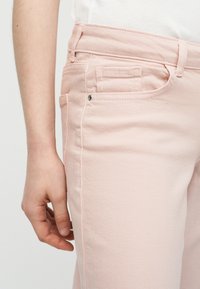 Κοντινό πλάνο αριστερής παλάμης ενός ατόμου που ακουμπάει δίπλα σε ανοιχτό ροζ τζιν με εμφανές μπροστινό τσέπη και θηλιές ζώνης.