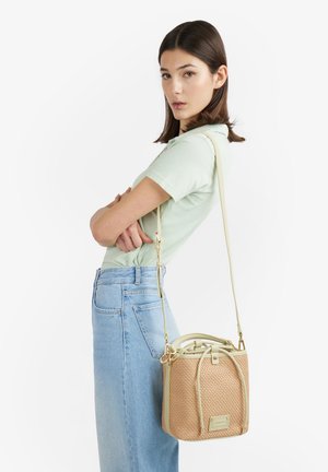 Donna con polo verde chiaro e jeans blu porta una borsa secchiello beige intrecciata con tracolla lunga sulla spalla, in piedi di profilo con le braccia incrociate.