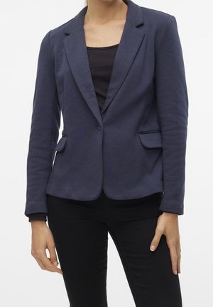Femme portant un blazer bleu marine cintré sur un haut noir et un pantalon noir, debout avec les bras détendus le long du corps sur un fond blanc.