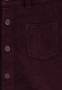 Bordeauxfarbener Cordstoff mit vertikalem Rippenmuster, vorderen Knöpfen und einer einzelnen Brusttasche. Die Textur ist weich und leicht erhaben.