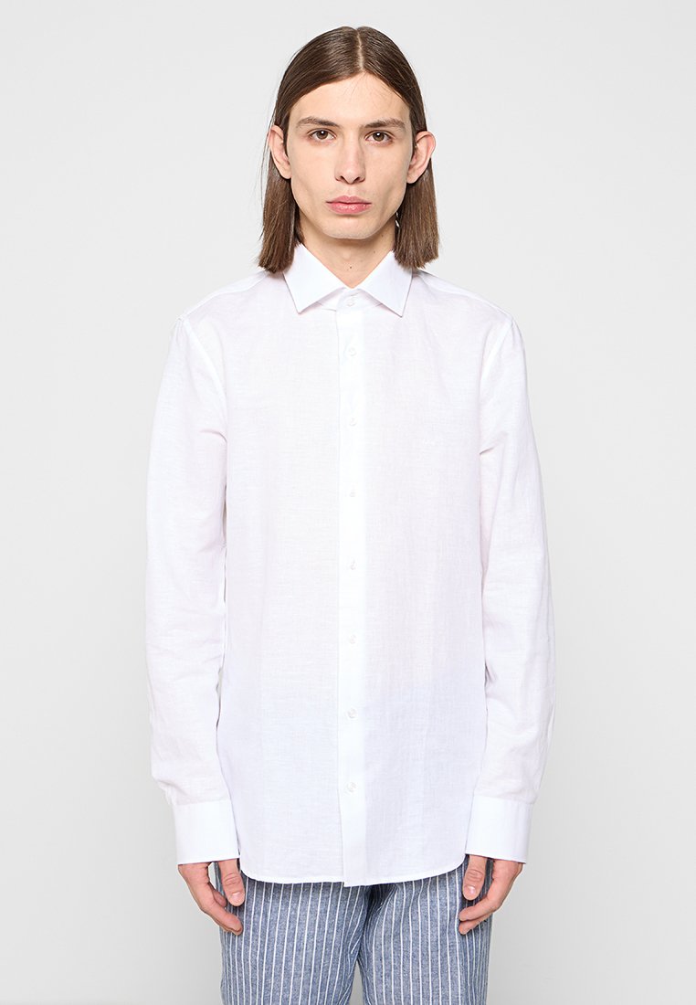 Młoda osoba z długimi brązowymi włosami, ubrana w gładką białą koszulę z guzikami oraz niebieskie paskowane spodnie, stojąca na tle jasnoszarym.