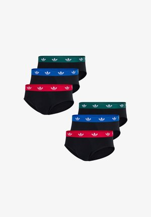 Skjorteløs mand iført sorte underbukser med hvide trefoil-logoer på taljebåndet, stående mod en ensfarvet lys baggrund.