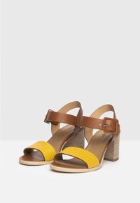 Sandalias de tacón de cuero marrón con una correa de ante amarilla, punta descubierta y un chunky tacón de bloque a rayas de color claro. Hebilla ajustable en el tobillo.