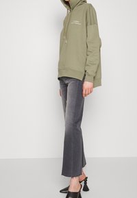 Sudadera con capucha de color verde oliva con un logo en el frente, combinada con jeans acampanados grises con dobladillos deshilachados y mule negras con borlas decorativas.