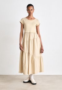 Beige gelaagde midi-jurk met korte mouwen, katoenen stof, ronde halslijn, gerimpelde taille en subtiele stikdetails, gedragen met crèmekleurige loafers.